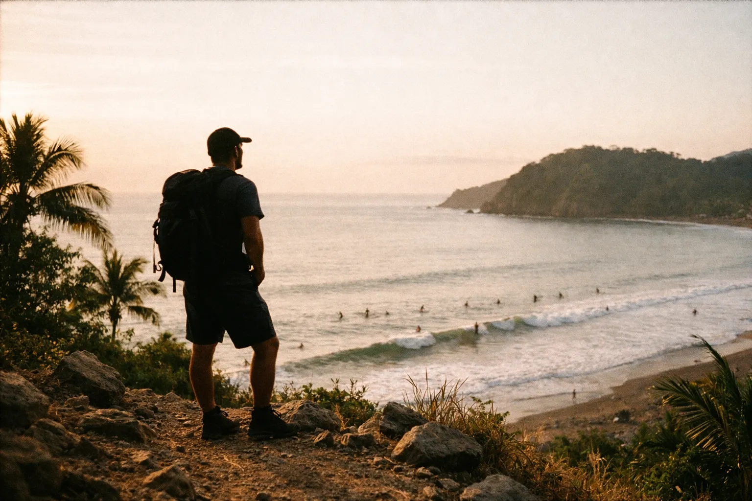 Hiker in silhouette overlooking Jaco coastline at golden hour; distant surfers