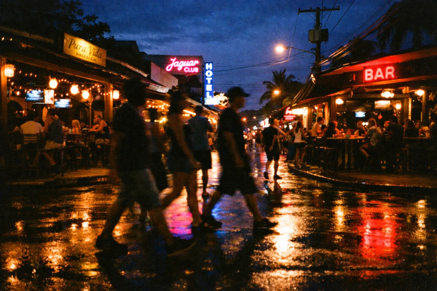 Street scene in Jaco at night with silhouettes strolling and warm streetlights, blurred neon signs