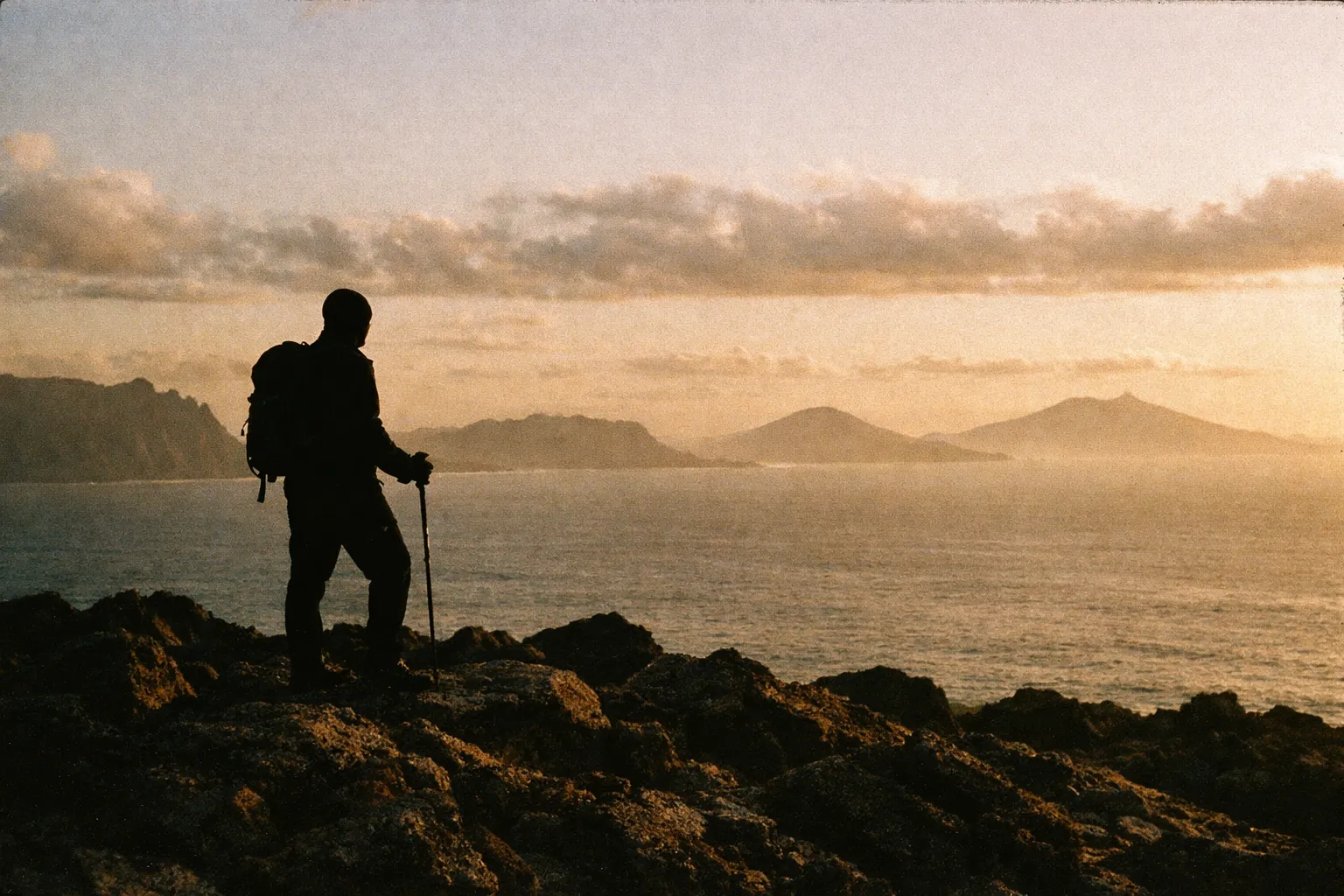 Silhouette of a lone hiker on a windswept overlook with distant island silhouettes on the horizon.