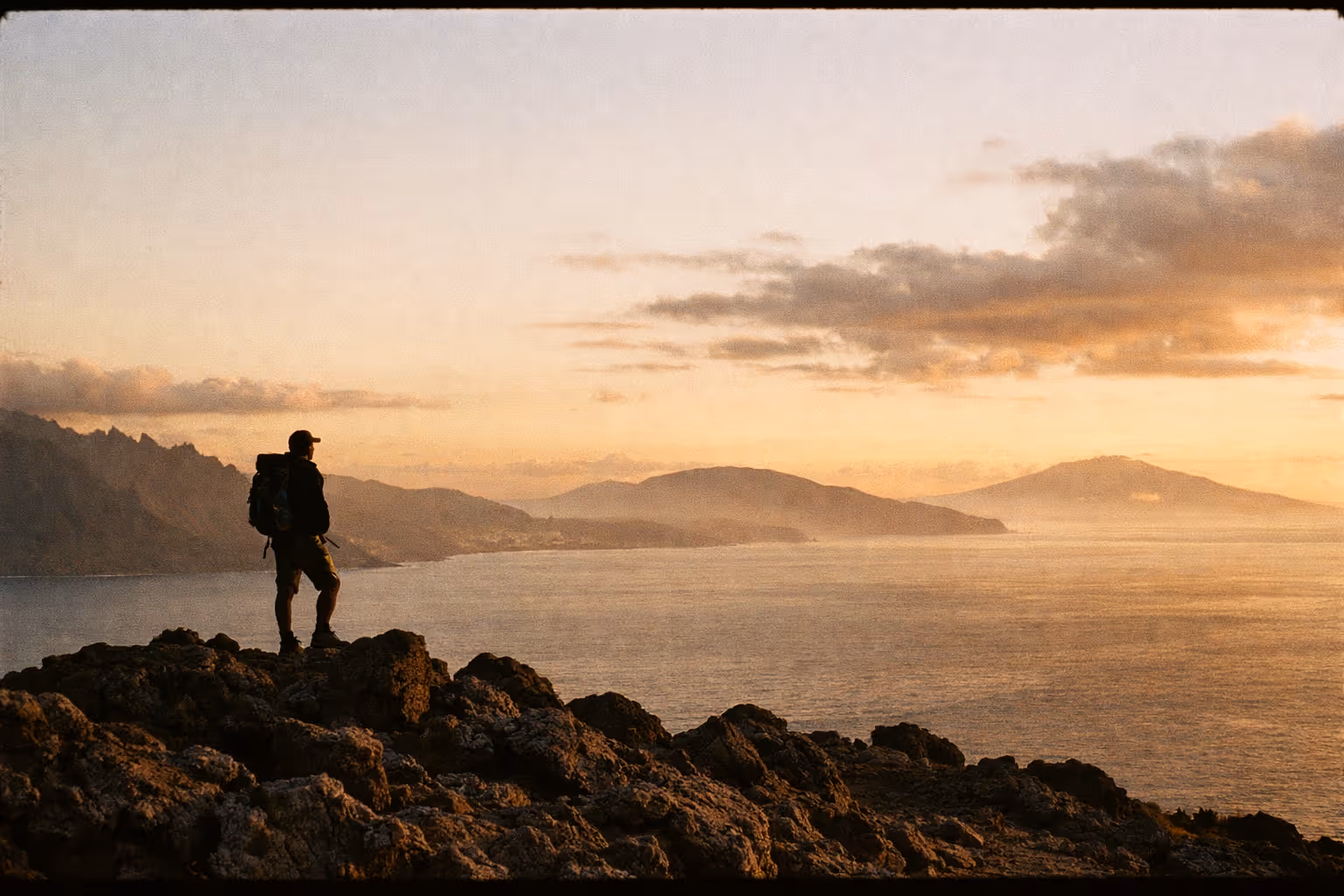 Silhouette of a lone hiker above Hawaii's wild landscape at golden hour, with Kauaʻi, Oʻahu, and Maui on the horizon.