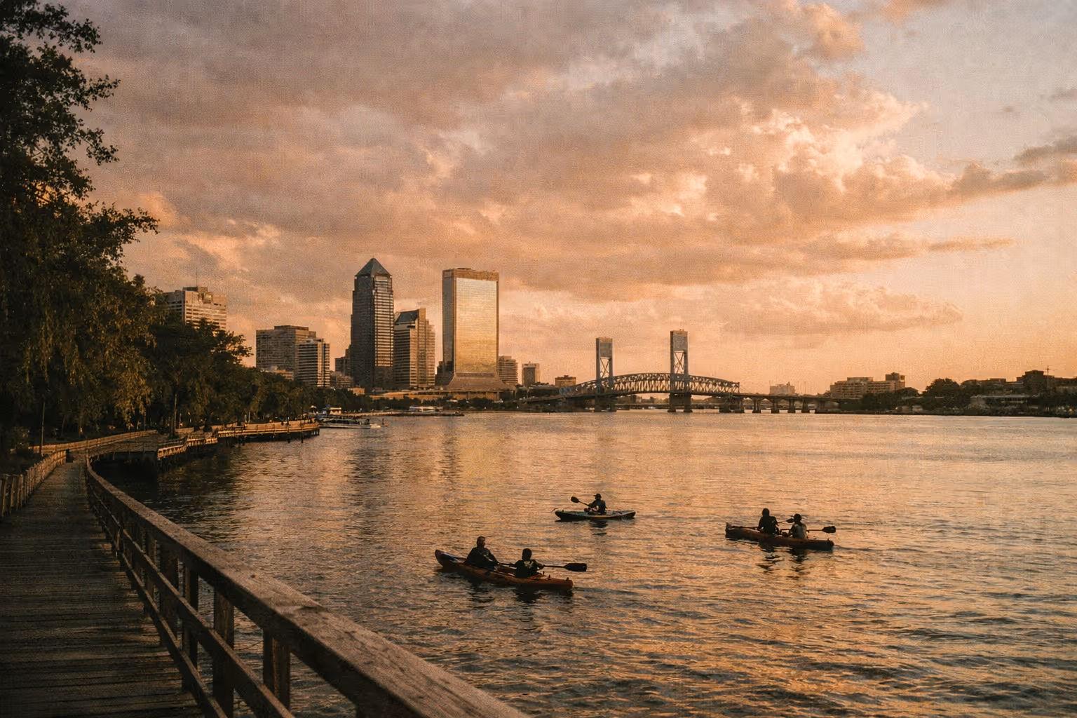Golden-hour wide shot of Jacksonville’s riverfront with silhouettes of kayakers on the St. Johns River, park trees in the foreground, and the downtown skyline in the distance.